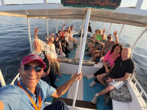       Cheerful tour group waves from an open boat cruising on calm Nile waters during golden afternoon light.
  