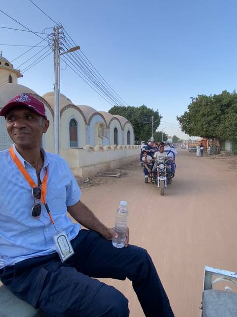       Guide looks on as a crowded motorbike taxi carries locals down a dusty village street beside distinctive Nubian architecture.
  