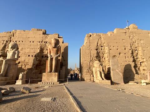       Visitors walk through the colossal gateway flanked by statues at Luxor Temple under a clear sky.
  