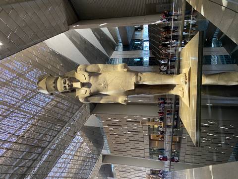       A colossal ancient Egyptian statue stands inside a modern museum atrium with visitors gathered below.
  