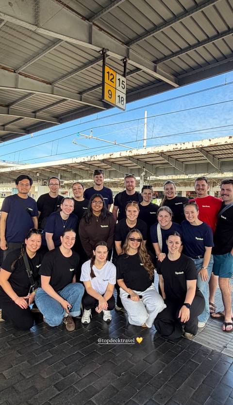       Large travel group posing for a photo at a train station platform under overhead wires.
  