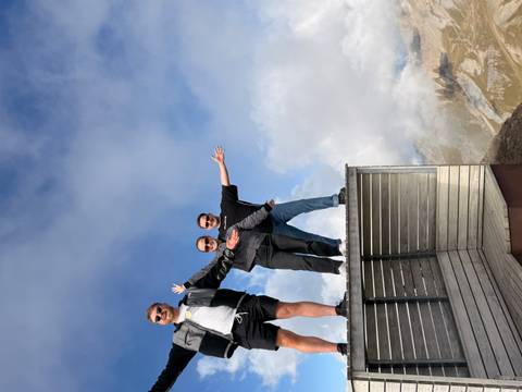       Three friends pose with arms outstretched on a mountaintop viewing deck above the clouds.
  