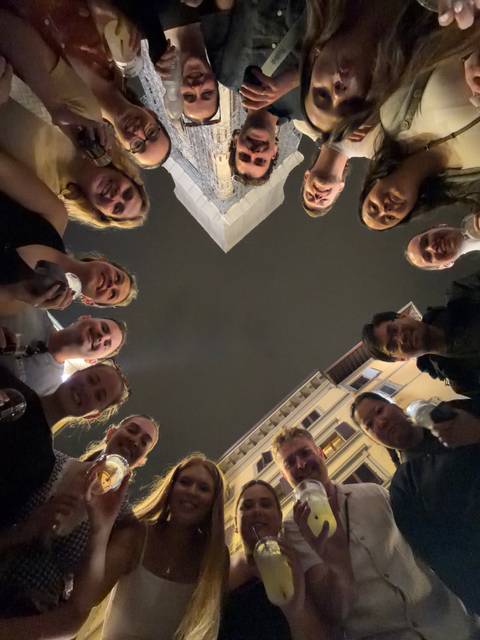       Night-time selfie taken from below with a circle of smiling friends holding drinks, open sky and surrounding buildings visible.
  