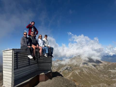       Travellers sitting on a wooden viewing platform high in the Alps with dramatic clouds and mountain peaks behind.
  
