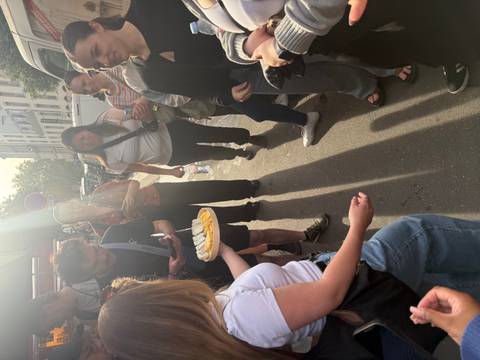      Group gathered on a Parisian street sampling cheese under bright afternoon sun.
  