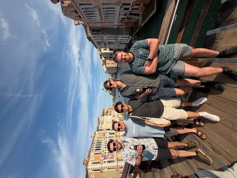       Five friends posing on a wooden bridge over Venice's Grand Canal with historic palaces and blue sky.
  