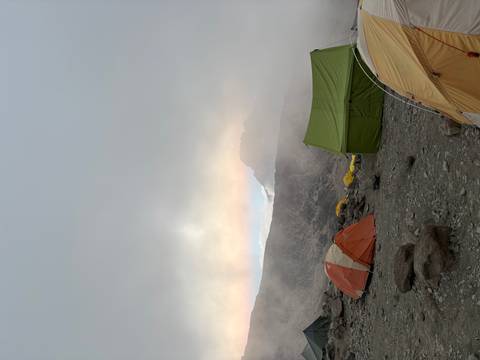       Tents set up on a rocky alpine campsite beneath swirling mist and faint mountain silhouette.
  