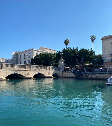       Historic stone bridge spans a turquoise canal lined with trees and cafés in Ortigia.
  