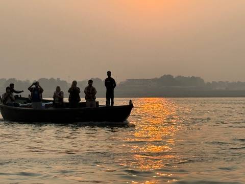       Silhouetted passengers stand in a motorboat on a river reflecting the orange glow of sunrise haze.
  