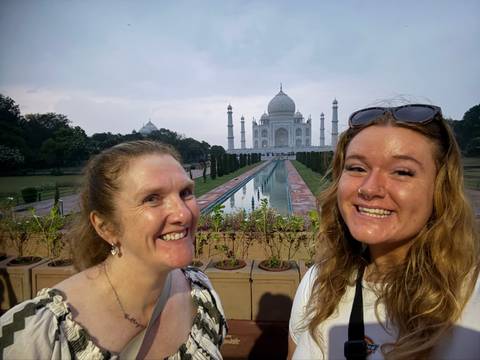       Two travellers smile in a selfie with the Taj Mahal and reflecting pool perfectly centered behind them.
  