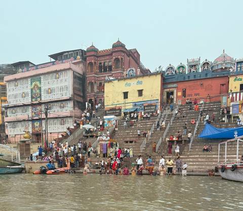       Crowds gather on the broad stone steps of a riverside ghat lined with brightly painted temples and houses in India.
  