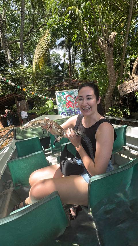       A woman proudly holds a small crocodile while standing on a boat surrounded by tropical greenery.
  