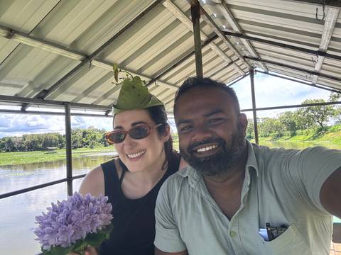       Two friends smile for a selfie on an open boat gliding across a calm lake under a leafy roof.
  