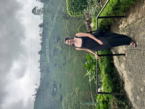       A visitor smiles before misty tea-covered hills with a large LABOOKELLIE sign in Sri Lanka’s highlands.
  