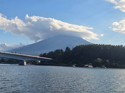       Clouds cloak the summit of Mount Fuji rising above a tranquil lake crossed by a modern bridge.
  