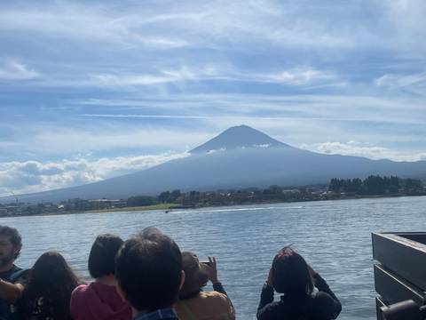       Mount Fuji rises above a lakeside town while tourists view the scene from a boat.
  