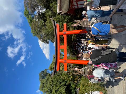       Crowds walk under the vermilion torii gate at Fushimi Inari Shrine on a sunny day.
  