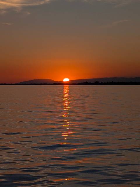       Serene sunset with the sun dipping behind distant mountains and its reflection stretching over a lake.
  