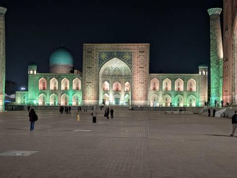       Registan square in Samarkand illuminated in green and gold lights at night with visitors walking across the plaza.
  