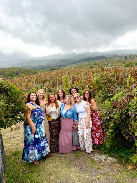       Group of smiling women posing among lush vineyard rows with misty mountains behind.
  