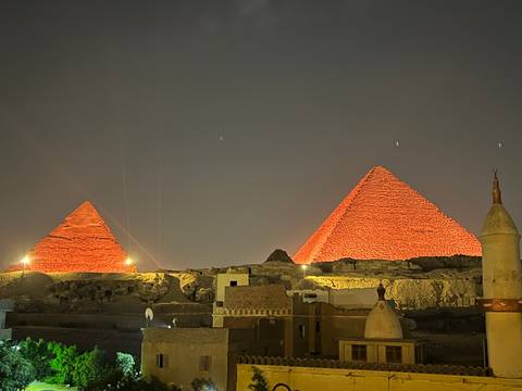       The Great Pyramids of Giza illuminated in red lights at night viewed over village rooftops.
  