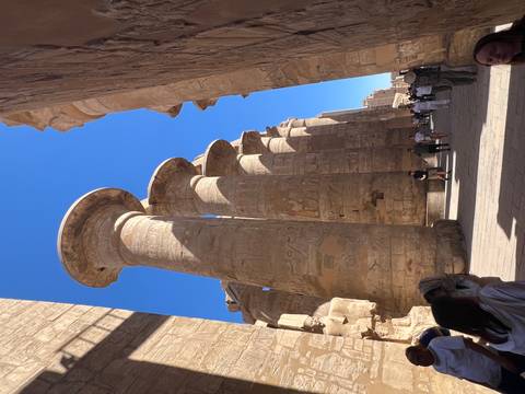       Immense sandstone columns of Karnak’s Hypostyle Hall towering over visitors beneath a bright blue sky.
  