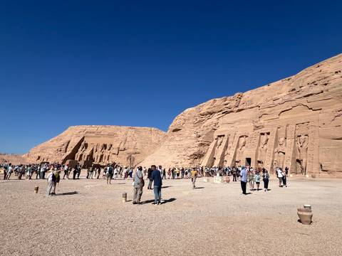       Wide view of Abu Simbel temples carved into sandstone cliff with crowds gathered outside on a clear day.
  