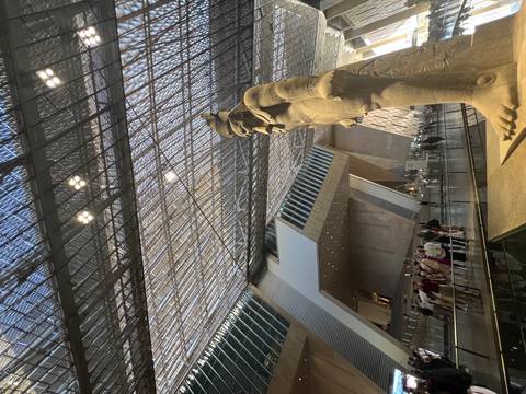       Interior of modern Grand Egyptian Museum with angular architecture and large ancient statue in foreground.
  