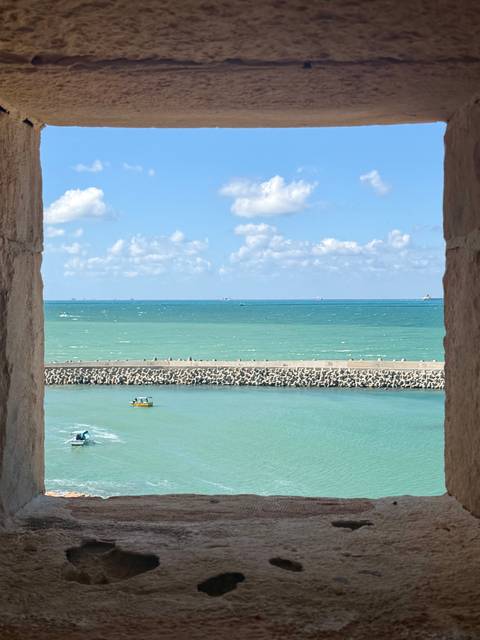       Turquoise Mediterranean waters and small boats framed by sandstone battlement window.
  
