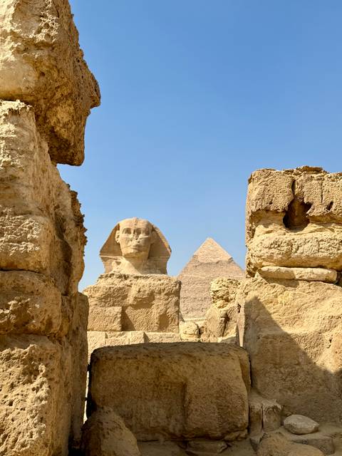      The Great Sphinx of Giza framed between weathered limestone blocks with a pyramid rising behind against clear sky.
  