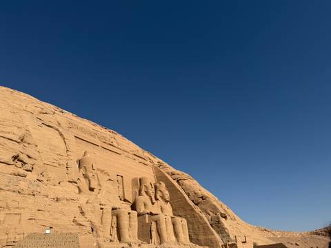      Colossal seated statues of Abu Simbel carved into sandstone cliff under cloudless deep blue sky.
  