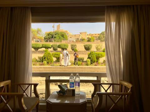       View through open cabin window of Nile cruise ship toward shore where two traditionally dressed locals stand among manicured gardens.
  