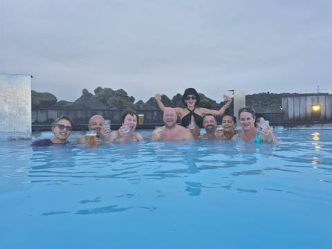       Happy bathers toast drinks while soaking in milky blue geothermal water surrounded by lava rocks.
  