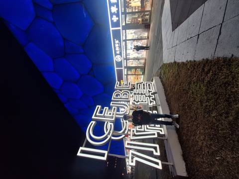       Young traveller posing in front of Beijing’s blue-lit Water Cube with bright ICE CUBE neon sign at night.
  
