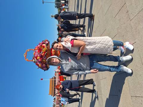       Mother and son smiling in Tiananmen Square under clear blue sky with giant flower basket decoration behind.
  