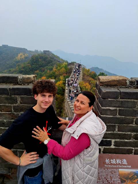       Two travellers posing on a tower of the Great Wall with autumn foliage and a long line of visitors stretching into the hills.
  