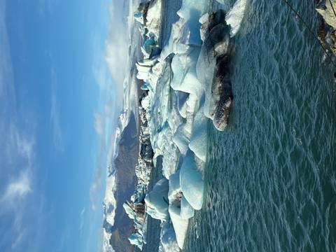       Expansive view of turquoise water dotted with many pale blue icebergs and snow-capped mountains behind.
  