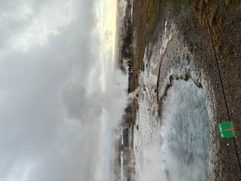       Steaming geothermal pool and erupting geyser plume rising into a cloudy sky.
  