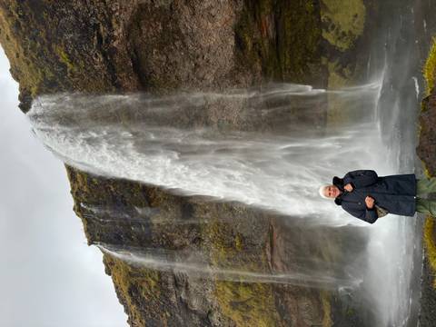       Elderly traveler standing in front of a tall, narrow waterfall cascading from a mossy cliff.
  