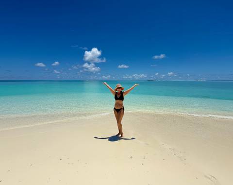       Woman in a black bikini poses joyfully on a pristine white-sand beach with clear aquamarine water.
  