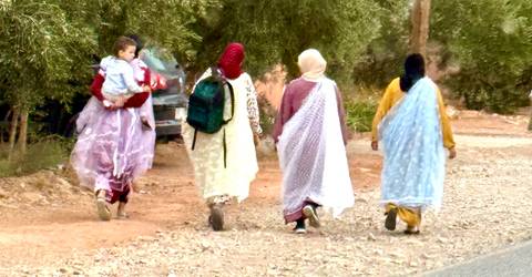       Women in colorful traditional garments walk along a dusty rural track, one carrying a child.
  