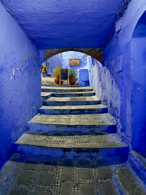       Stone staircase between vivid blue walls in Chefchaouen, with potted plants and a small restaurant sign.
  