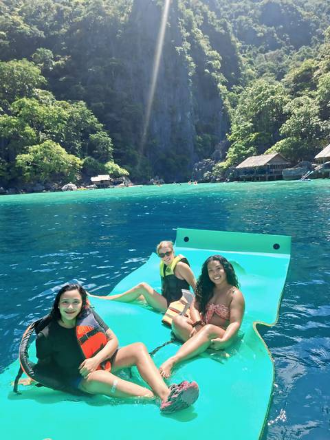       Three women lounge on a bright green floating mat in crystal-clear lagoon waters backed by towering limestone cliffs and jungle.
  