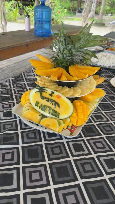       Tiered fruit tray with a carved melon reading 'I ❤ Port Barton' surrounded by pineapple and mango slices on a patterned cloth.
  