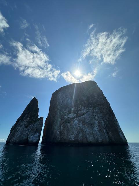 Large rock formation with the sun shining behind it, surrounded by blue sky and sea.