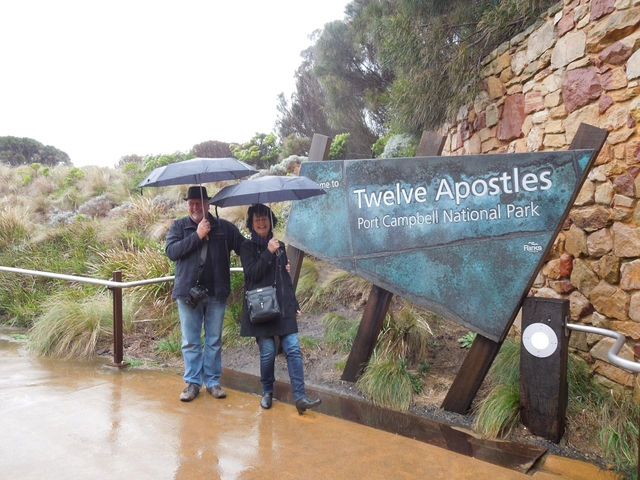       Two people with umbrellas in front of a sign for Twelve Apostles Port Campbell National Park.
  