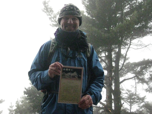       Smiling person holding a certificate in a forest setting.
  