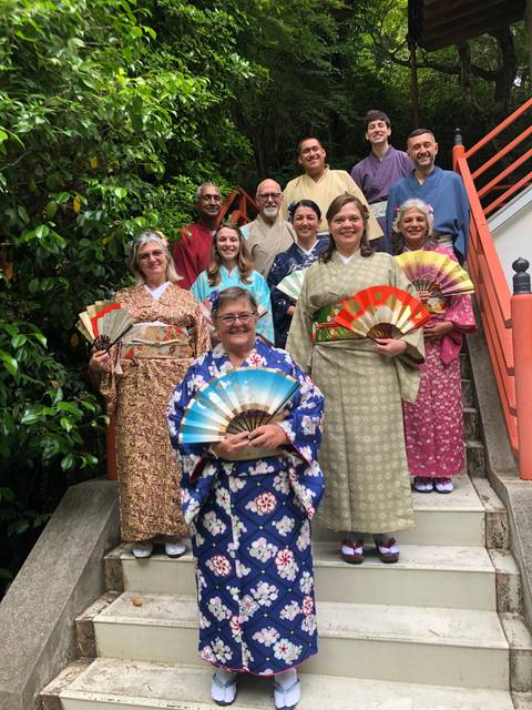       Group dressed in traditional kimonos standing on stairs with greenery surrounding.
  