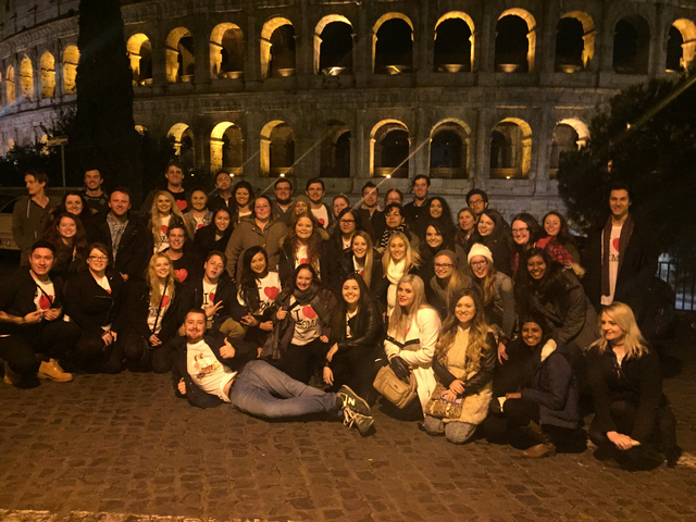 Large group posing in front of a historical amphitheater at night.