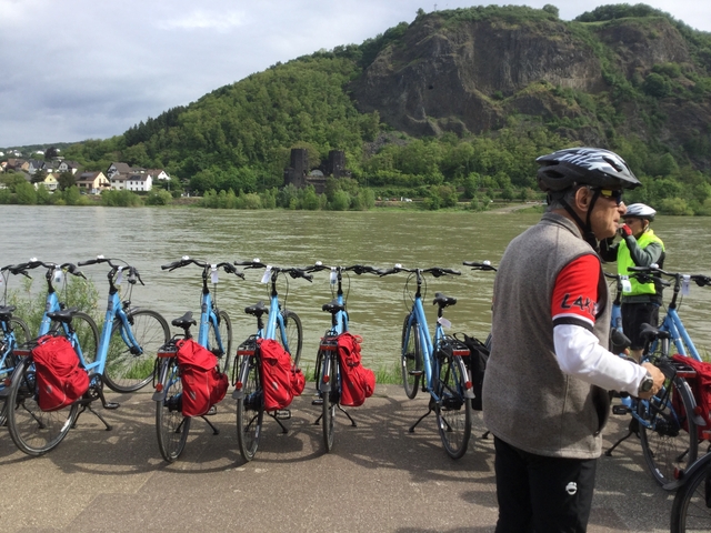 Cyclists with bicycles near a river, small village in the background.